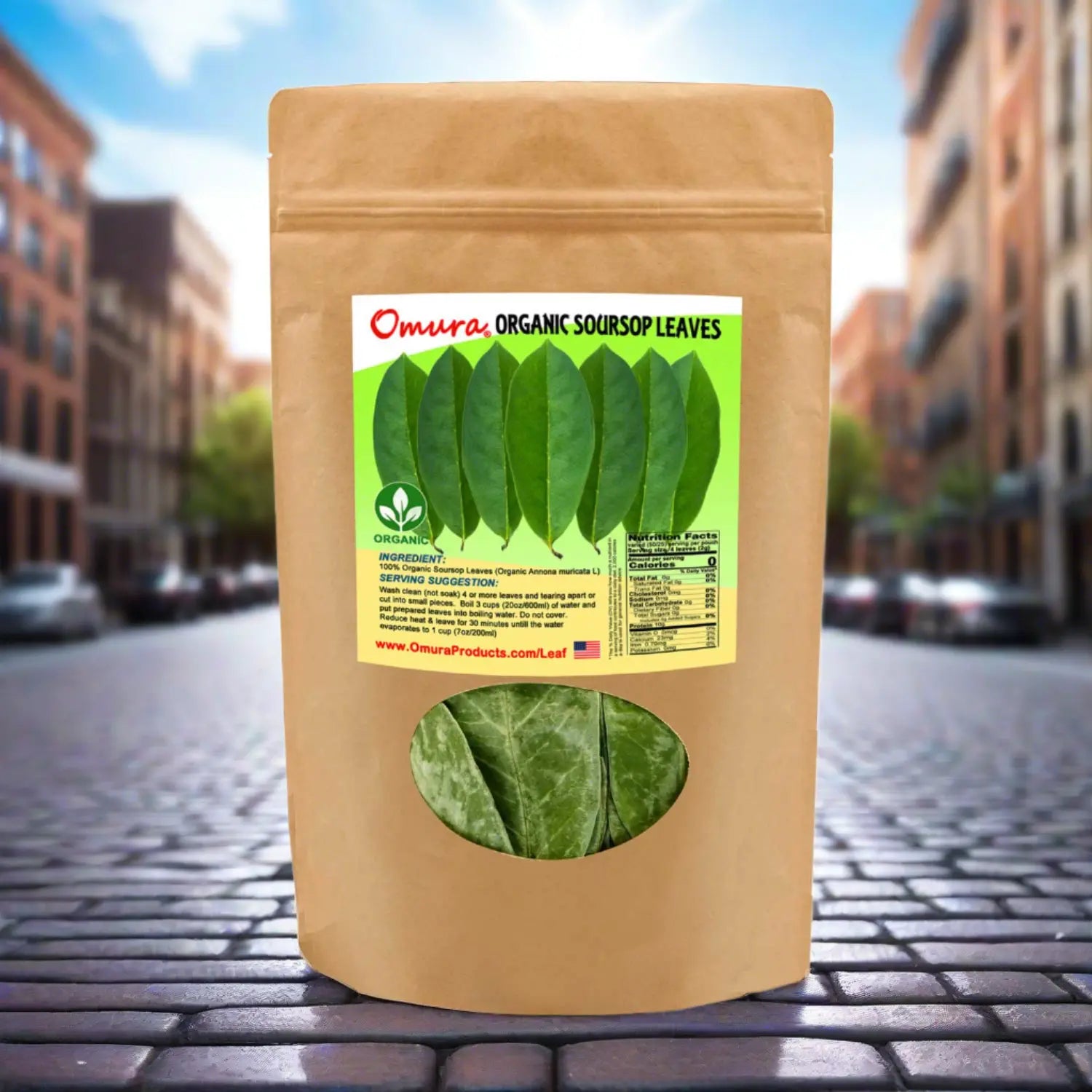 Packaging of Omura organic soursop leaves with a clear window showing the green leaves inside. The package is placed on a cobblestone street with buildings in the background.